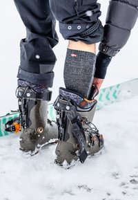 Botas de esquí en gris oscuro con acentos naranjas, con un collar alto y hebillas ajustables. Fondo cubierto de nieve con un esquí bordeado de verde.