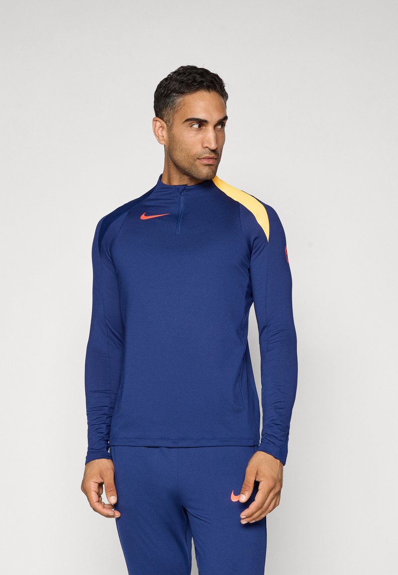 Marineblå langermet treningsgenser med kvart glidelås, gul skulderdetalj, rød Nike-logo. Glatt, lett stoff.