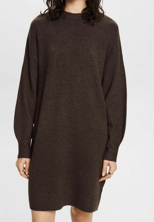 Femme portant une robe-pull en maille marron foncé à manches longues, avec un col rond et des poignets ajustés, sur fond blanc.