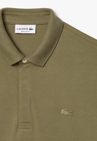 Polo di colore verde oliva chiaro in cotone tessuto, con colletto a due bottoni e un piccolo logo di coccodrillo ricamato sul petto.