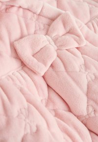 Couverture en peluche rose clair avec un design matelassé. Comprend un grand nœud en tissu en accent. Surface douce et texturée avec des motifs floraux subtils.