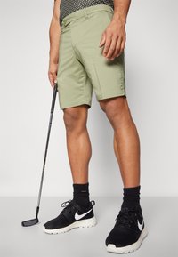 Kaki golfa īsās bikses ar priekšējiem un aizmugurējiem kabatām, apvienotas ar melnām Nike kurpēm un melnām zeķēm, turēt golfa nūju. Tekstūr�ēts dizains.