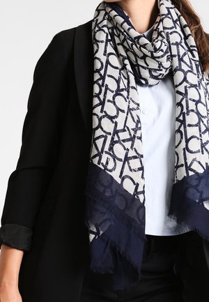 Femme portant un blazer noir et une chemise blanche à boutons, accessoirisée d'une longue écharpe blanche ornée de grosses lettres noires et de bordures bleu marine foncé.
