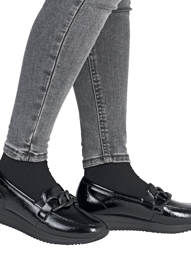 Black patent leather loafers with a round toe, featuring a black chain accent on the front, worn with gray skinny jeans and black socks.