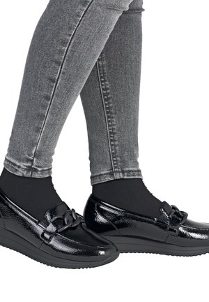 Personne portant des mocassins en cuir verni noir avec un détail de chaîne décorative et un jean skinny gris sur fond blanc.