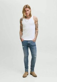 Uomo con capelli lunghi e tatuaggi indossa una canotta bianca, jeans skinny strappati, stivali beige e una collana di catena argentata, in piedi con le mani nelle tasche.