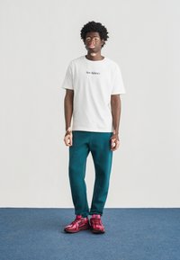 Άσπρο κοντομάνικο μπλουζάκι με το λογότυπο «New Balance», τιρκουάζ παντελόνι φούτερ και ροζ αθλητικά παπούτσια με μαύρες λεπτομέρειες, στέκεται σε μπλε χαλί.