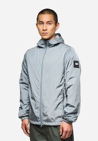 Giacca con cappuccio grigio chiaro realizzata in tessuto ripstop, caratterizzata da una cerniera frontale e polsini elastici. Fodera nera sulla manica sinistra.