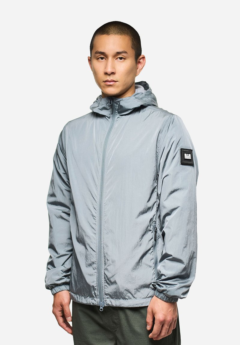 Giacca con cappuccio grigio chiaro realizzata in tessuto ripstop, caratterizzata da una cerniera frontale e polsini elastici. Fodera nera sulla manica sinistra.