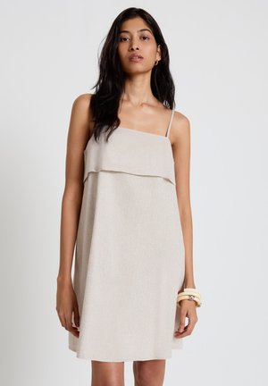 Donna con capelli lunghi e scuri che indossa un vestito beige senza maniche e braccialetti bianchi, in piedi contro uno sfondo chiaro e uniforme.