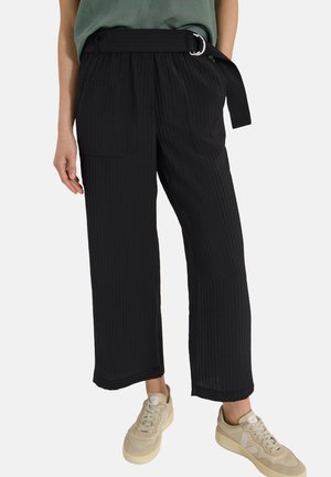 Femme portant un pantalon noir à jambes larges avec de grandes poches avant et une ceinture noire, associé à des baskets beige et un haut vert.
