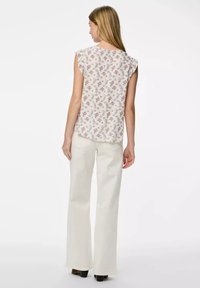 Top senza maniche floreale color crema con fiori viola e rosa; orlo arrotondato; abbinato a pantaloni chiari a zampa d'elefante; sfondo neutro.