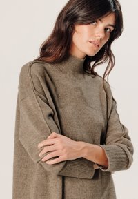 Übergroßer, olivgrüner, gestrickter Pullover mit hohem Kragen, gerippten Bündchen und glatter Textur, präsentiert mit überschnittenen Schultern und dezenten Details.