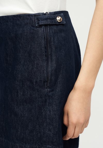Κοντινό πλάνο ενός ατόμου που φοράει σκουρόχρωμη μπλε τζιν φούστα με πλευρικό φερμουάρ και λεπτομέρεια με κουμπί, σε συνδυασμό με λευκό τοπ.