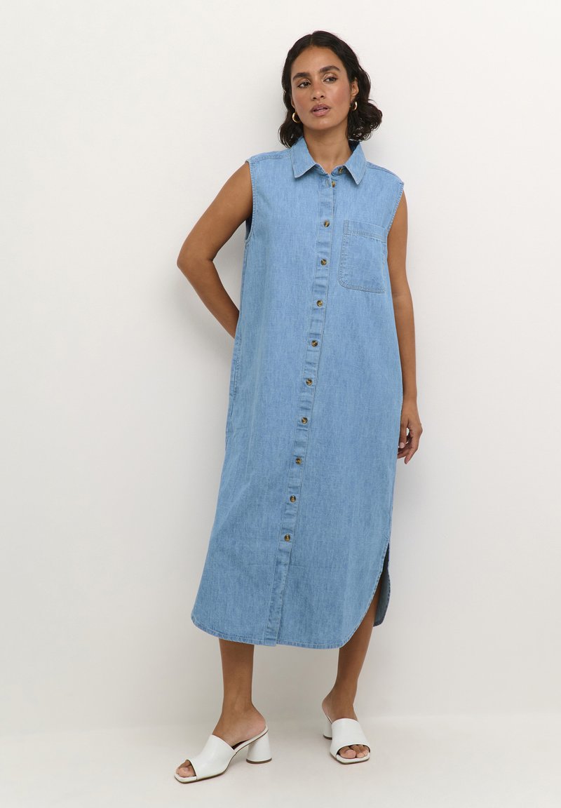 Vestido de mezclilla sin mangas de color azul claro, con un bolsillo en el pecho y aberturas laterales. Combinado con unos zuecos blancos de tacón.