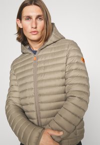 Beige puffer jacket with horizontal seams and a hood. Features a front zipper, orange zipper pull, and a round logo patch on the sleeve.