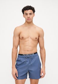 Vīriešu zili peldbikini ar melnu elastīgu jostasvietu, uz kuras ir "CALVIN KLEIN" logo un regulējams šnērs. Gluda, viegla auduma.