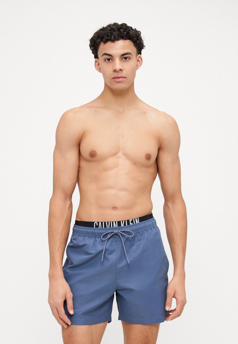 Férfi kék fürdőnadrág fekete elasztikus derékkal, amelyen a "CALVIN KLEIN" logó látható, állítható zsinórral. Simább, könnyű anyag.