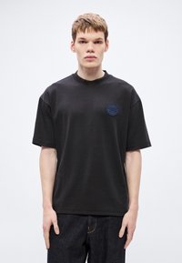 Μαύρο βαμβακερό T-shirt με άνετη εφαρμογή, στρογγυλή λαιμόκοψη και κοντά μανίκια· διαθέτει στρογγυλή μπλε κεντημένη επιγραφή στο αριστερό στήθος.