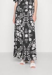 Czarna spódnica z białym kwiatowym i liściastym wzorem, o luźnym kroju i szerokim rozcięciu. Połączona z białymi, teksturowanymi sandałami na otwartym palcu.