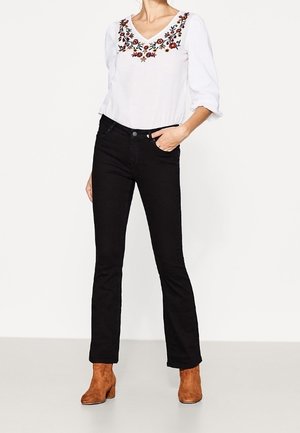 Weißes Blusenshirt mit floralem Stickereimuster am Halsauschnitt, kombiniert mit schwarzen, figurbetonten Jeans und braunen Stiefeletten. Glattes Material und schlanke Form.
