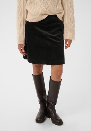 Personne portant un pull beige torsadé, une jupe en velours côtelé noire et des bottes en cuir marron foncé jusqu'aux genoux, debout sur un fond uni.