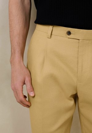 Male wearing beige trousers with a button closure and a black knit top, arm relaxed by side against a neutral background.