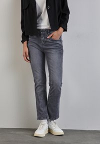 Persona con jeans grises desgastados, zapatillas blancas y chaqueta negra, con una mano en el bolsillo, de pie contra una pared y un suelo claros.