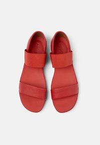 Red sandals featuring a smooth leather upper with wide elastic straps and a flat sole, designed for comfort and ease of wear.