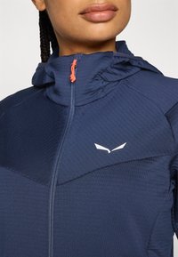 Jūrinės tamsiai mėlynos spalvos zip-up striukė su vertikaliu apykakle, tekstūruota medžiaga, balta logotipo detalė ir oranžinis užtrauktuko traukiklis. Išsiskiria prigludusia forma.