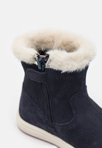 Bottine en suede marine avec un col doublé en fourrure, fermeture à glissière latérale et semelle en caoutchouc blanche. Elle présente une texture douce et des coutures contrastées.