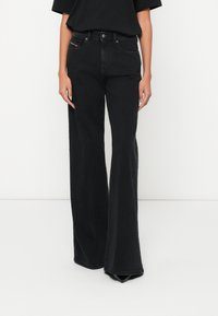 Γυναίκα που φοράει ψηλόμεσο μαύρο τζιν καμπάνα με πέντε τσέπες και μαύρα μυτερά παπούτσια, σε λευκό φόντο.