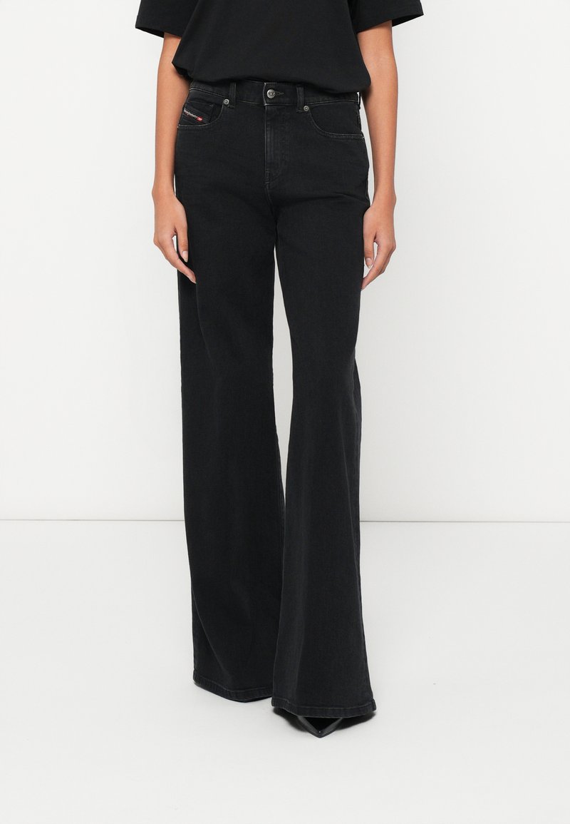 Γυναίκα που φοράει ψηλόμεσο μαύρο τζιν καμπάνα με πέντε τσέπες και μαύρα μυτερά παπούτσια, σε λευκό φόντο.