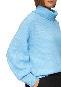 Lichtblauwe gebreide trui met een hoge col, geribbelde structuur en oversized mouwen. Het materiaal lijkt zacht en comfortabel te zijn.
