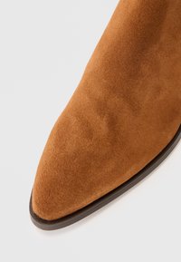 Brązowy zamszowy botek sięgający kostki z ostrym noskiem, gładką powierzchnią i kontrastującą czarną podeszwą. Brak widocznych elementów metalowych lub wzorów.