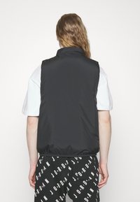 Crni bezrukavnik izrađen od glatkog materijala, s visokim ovratnikom i elastičnom donjom ivicom, nosi se preko bijele košulje i uzorčanih crnih hlača.