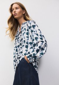 Biała bluzka z długim rękawem w granatowy abstrakcyjny wzór kratki, o luźnym kroju, marszczonym dekolcie i lekko teksturowanej tkaninie.