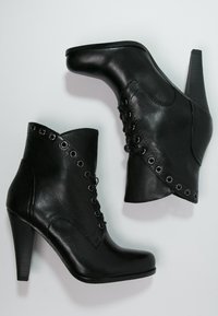 Botas de cuero negras hasta el tobillo con punta afilada, tacón alto, parte delantera con cordones y ojales metálicos decorativos en el lateral. Textura suave.