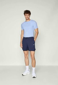 Lyseblå, tettsittende skjorte med kort erm og teksturerte sider, sammen med mørk marineblå treningsshorts med elastisk midje og logo. Hvite sneakers.
