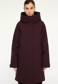 Vrouw gekleed in een lange, donkerpaarse winterjas met een hoge kraag en capuchon, staande met de handen langs haar zij tegen een effen achtergrond.