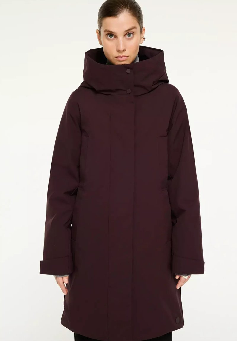 Vrouw gekleed in een lange, donkerpaarse winterjas met een hoge kraag en capuchon, staande met de handen langs haar zij tegen een effen achtergrond.