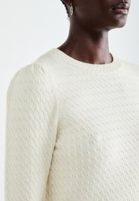 Pull en crème clair avec une texture, présentant un subtil motif géométrique, un col rond et des manches bouffantes.