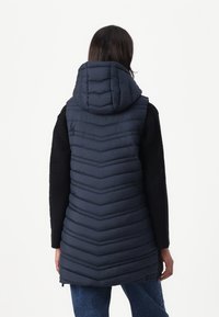 Donkerblauwe, lange gewatteerde vest met een chevronpatroon, verstelbare capuchon en gestructureerde zwarte gebreide mouwen. Ritssluiting aan de voorkant.
