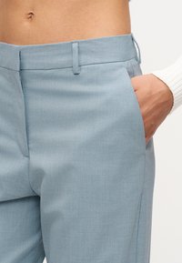 Το χέρι ενός ατόμου ακουμπάει μέσα στην δεξιά τσέπη ανοιχτόχ ρωμων παντελονιών, εμφανίζοντας τη μέση και μέρος ενός λευκού μακρυμάνικου τοπ.