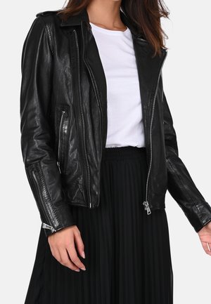 Kobieta ubrana w czarną skórzaną kurtkę z srebrnymi zamkami błyskawicznymi, białą koszulkę i czarną plisowaną spódnicę, stojąca na jednolitym tle.