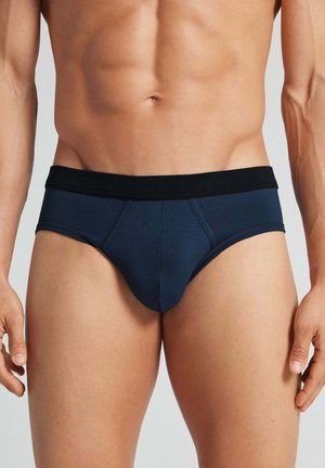 Mandlig torso iført mørkeblå trusser med sort elastik mod en ensfarvet baggrund.