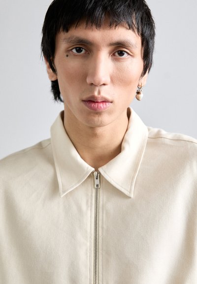 Veste zippée beige clair avec un col pointu, confectionnée dans un tissu lisse, dotée d'une fermeture éclair argentée et de détails texturés subtils.