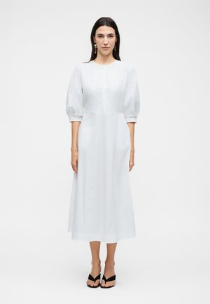 Žena s dlhými tmavými vlasmi oblečená v bielych midi šatách s nadýchanými rukávmi a čiernych sandáloch na podpätku stojaca pred jednofarebným pozadím.