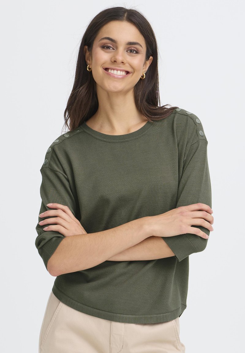Femme souriante aux longs cheveux bruns portant des boucles d'oreilles en or, un haut olive à manches boutonnées et un pantalon beige, les bras croisés.
