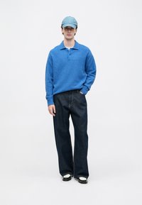 Jovem com um boné azul, camisa de polo de manga longa texturizada azul, jeans largos escuros e sapatos pretos e brancos, parado com uma mão no bolso.